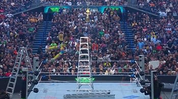 A wrestler performs an aerial maneuver off a tall ladder during a dramatic moment in the ring, with a large crowd cheering in the background. The object of contention is suspended above, adding tension to the scene.