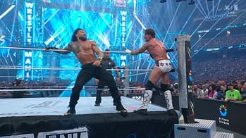 In a wrestling match, a muscular athlete in black pants and no shirt is facing off against another wrestler wearing white tights with black stars. The energetic atmosphere is heightened by bright blue lights and a large crowd in the background.