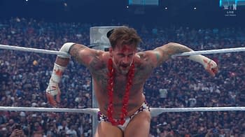 A muscular wrestler with tattoos and a bloodied face celebrates in the ring, wearing a flower lei and white shorts, with fans visible in the background.