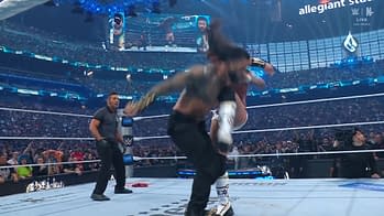 A wrestling match at WrestleMania features a wrestler executing a move on another, with a referee looking on. A large crowd is visible in the background under colorful lights.