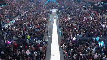 A large crowd gathers at a wrestling event, with colorful lights illuminating the scene. A long white entrance ramp leads toward a wrestling ring, highlighting the excitement of the occasion.