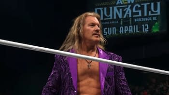 A wrestler wearing a purple sequined jacket stands in a wrestling ring, looking toward the audience. Behind him is a large banner promoting an upcoming AEW Dynamite event.