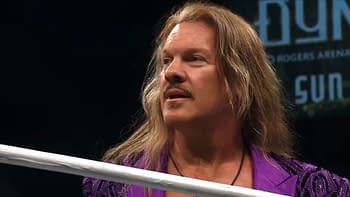 Professional wrestler Chris Jericho, wearing a purple jacket, stands in a wrestling ring, looking off to the side with a thoughtful expression. The background is dimly lit with signs for AEW Dynamite.