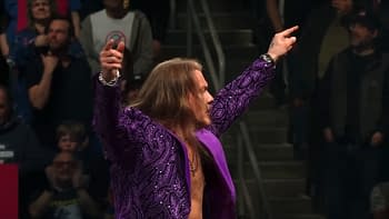 A professional wrestler with long hair raises his arms triumphantly while wearing a flashy purple jacket, amidst a cheering crowd. The atmosphere is electric, reflecting the excitement of a wrestling event.