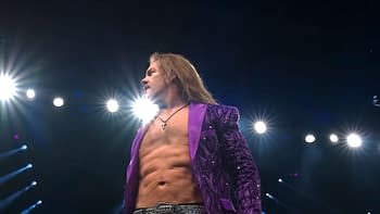 A muscular wrestler with long hair is wearing a shiny purple jacket, standing confidently under bright stage lights. He appears to be engaged with the audience.