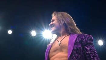 A muscular man with long hair stands under bright lights, smiling confidently. He is wearing a stylish purple jacket, showcasing a mix of flamboyance and athleticism.