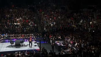 A wrestling ring is set up in front of a massive crowd, with fans eagerly watching a performer in the AEW arena. The performer is in the ring, surrounded by AEW branding and equipment.