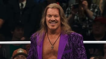 A man with long hair and a mustache wears a vibrant purple suit with intricate designs, smiling confidently. He stands in a wrestling ring with a cheering crowd in the background.