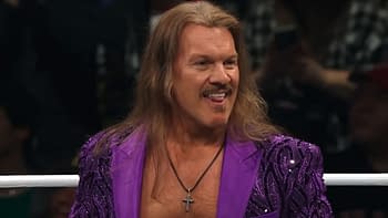 A man with long hair and a mustache wears a vibrant purple jacket adorned with sequins, smiling cheekily as he sticks out his tongue. He is in a wrestling ring, surrounded by a captivated audience.