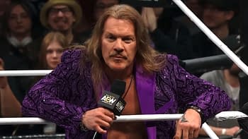 A wrestler in a purple jacket leans on the ropes of a wrestling ring, holding a microphone. He has long hair and a distinctive mustache, with the audience visible in the background.