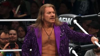 A male wrestler with long hair wearing a purple sequined jacket stands in a wrestling ring. He appears confident, engaging with the audience in the background.