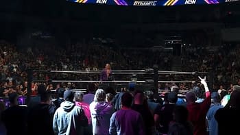 A crowd at a wrestling event reacts as Chris Jericho makes a dramatic return to AEW on Dynamite, wearing a purple jacket and standing in the ring. The audience shows a mix of excitement and surprise, with fans visible in the foreground and AEW branding displayed above.