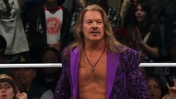 A male professional wrestler with long hair and a purple sequined jacket stands in a wrestling ring, facing the audience. He has a serious expression, and a crowd can be seen in the background.