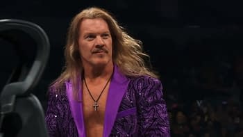 A man with long hair and a mustache wears a purple sequined jacket with a cross necklace, appearing in a wrestling ring setting. The crowd is blurred in the background.