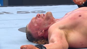 A wrestler lying on the mat, with facial expression indicating exhaustion or defeat. The scene captures the aftermath of a wrestling match, reflecting the physical intensity of the sport.