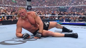 Brock Lesnar lies on the wrestling ring floor, appearing exhausted and defeated after a match, wearing black shorts and gloves. The background shows a large crowd of spectators in a wrestling arena.