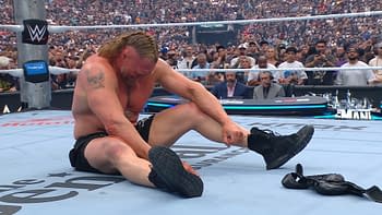 A wrestler, with long hair and a tattoo, sits on the wrestling mat, visibly in pain after a match, surrounded by a cheering crowd. The arena is filled with spectators capturing the moment on their phones.