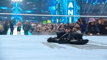 A discarded pair of wrestling boots lies on the mat in the foreground, while a crowd watches from the background of a wrestling event filled with bright lights and large screens.