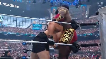 A wrestling match featuring Oba Femi in a colorful outfit overpowering Brock Lesnar, who is wearing black shorts. The background shows a large audience in a stadium filled with lights and screens.