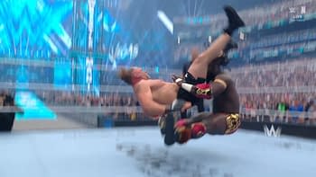 A wrestling match scene capturing Oba Femi performing a dramatic move, lifting Brock Lesnar upside down. The background shows a crowded arena with vibrant lighting effects.