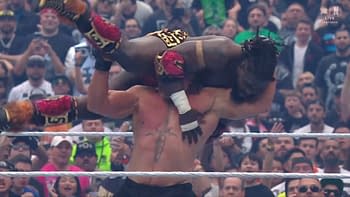 A wrestler with dark skin and colorful attire is being lifted above his opponent's head in a wrestling ring. The audience is visible in the background, showing various expressions of excitement.
