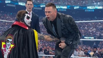 A performer dressed as a clown-like character with face paint and a red and black cape stands facing another character in a formal suit, who looks shocked. In the background, a wrestling ring and an audience are visible.