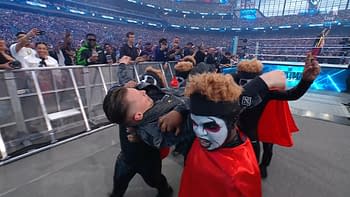 A group of performers, dressed in black and red attire with clown-like makeup, is in the midst of a wrestling-themed event. One performer is being lifted by another as they interact with a lively crowd in the background.