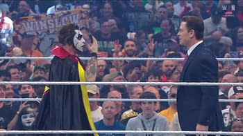 A performer dressed as a character with a painted face and a cape engages in a dramatic moment in a wrestling ring, surrounded by a cheering audience holding signs and capturing the moment with their phones.