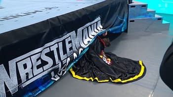 A person in a black cape with yellow trim is partially hiding under a wrestling ring banner. The scene is set at WrestleMania, indicated by the banner's logo.