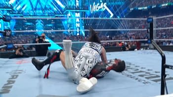 A wrestling match is taking place in a brightly lit arena, with one wrestler pinned down by another on the mat. The background features colorful lights and an audience filled with spectators.