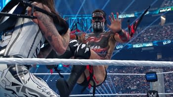 A professional wrestler with intricate face paint, featuring black, red, and white designs, is executing a kick in a wrestling ring. The scene captures an intense moment during a match at WrestleMania with a cheering audience visible in the background.