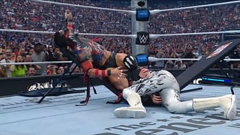 A dramatic wrestling match scene with one wrestler in a colorful outfit executing a move on another wrestler, who is lying on the mat. The audience in the background appears enthusiastic, surrounded by the wrestling ring.