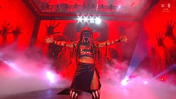 A wrestler with an elaborate costume and face paint stands with outstretched arms against a dramatic red backdrop, surrounded by fog and lighting effects.