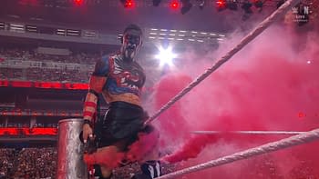 A wrestler with elaborate body paint stands in a wrestling ring surrounded by pink smoke, showcasing vibrant colors and an intense expression. The venue is filled with spectators in the background, illuminated by red arena lights.
