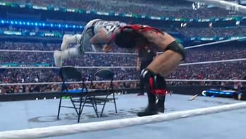 A dramatic wrestling moment featuring a performer in white gear leaping off a chair towards another wrestler in red and black attire, with an enthusiastic crowd visible in the background.
