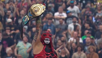 A masked wrestler in a vibrant red outfit triumphantly holds up a championship belt above their head in front of a cheering crowd. The background shows enthusiastic fans celebrating the moment.