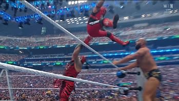A wrestler in red attire leaps off the ropes, poised mid-air, aiming to land on an opponent below during a high-stakes match in a crowded arena. The audience can be seen in the background, filled with excitement.