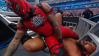 A wrestler in a red costume is on top of another wrestler due to a maneuver commonly seen in wrestling matches. The background shows an audience in a large arena during a wrestling event.