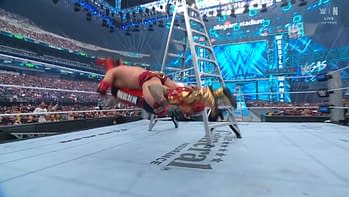 In a dramatic moment from a wrestling match, a wrestler in red gear performs a move while being propelled towards a ladder, with a large audience visible in the background under bright lights.
