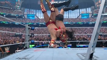 A dramatic moment in a wrestling match featuring two wrestlers in mid-air, with one performing a powerful move against the other in a large arena filled with spectators. The ring is surrounded by ropes and a ladder is visible nearby.