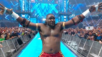 A muscular wrestler celebrates with arms raised, wearing red shorts and displaying a determined expression. A cheering crowd is visible in the background, with colorful signs and various fans.