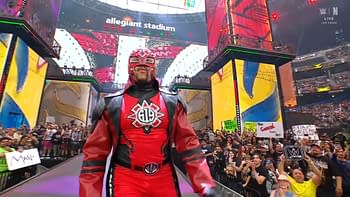 A wrestler dressed in a red and black suit with a dramatic mask walks through the crowd at Allegiant Stadium during a wrestling event, surrounded by enthusiastic fans holding signs.