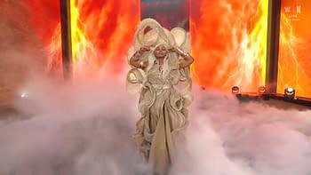 A female wrestler with long, flowing hair styled dramatically, dressed in a flowing golden outfit, poses confidently amidst fog and a backdrop of bright orange flames and lightning effects.
