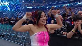 A woman in a colorful outfit with pink and white accents expresses excitement at a wrestling event, raising her arms in celebration. The crowd behind her is energetic and engaged in the moment.