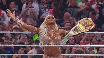 A wrestler celebrating in the ring with arms raised and holding a championship title. She is wearing a shimmering outfit and has long, blonde hair, while fans in the background are taking pictures.