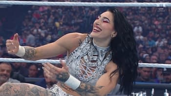 A happy woman with long dark hair and striking makeup smiles and gestures while wearing a shiny, studded outfit in a wrestling ring surrounded by cheering fans. Her tattoos are visible on her arms as she engages with the audience.