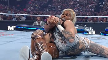 A female wrestler in a white outfit with tattoos applies a submission hold on her opponent in a wrestling ring at WrestleMania, with a crowd visible in the background.