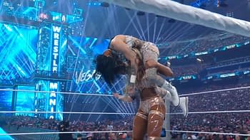 A wrestling match featuring a female wrestler lifting another wrestler in a dramatic move, surrounded by bright lights and a cheering crowd at a large arena. The event is WrestleMania, with branding visible in the background.