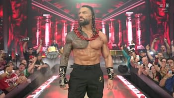 A muscular wrestler, Roman Reigns, stands confidently in a dramatic arena setting, wearing a red necklace and black attire. He faces a cheering crowd amidst vibrant lights and elaborate backdrop designs.