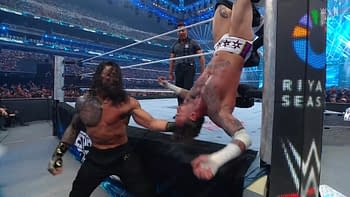 A dramatic moment in a wrestling match shows a muscular wrestler lifting an opponent upside down against the corner post, with a large cheering crowd in the background. The wrestler appears determined, while the opponent seems in a vulnerable position.